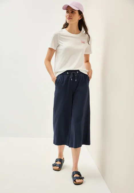 Frau mit weißem T-Shirt, blauer Culotte, Sandalen und lila Kappe.